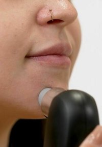 Dispositif de nettoyage facial noir avec un embout applicateur en silicone blanc, près d'une joue lisse et légèrement texturée. La peau semble saine et claire.