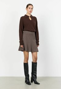 Suéter marrón de punto con cuello y detalle de abertura, combinado con una falda marrón de cuadros houndstooth. Botas de cuero negras hasta la rodilla.