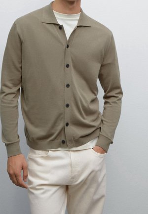Homme portant un cardigan taupe boutonné sur un t-shirt blanc avec un pantalon crème, main dans la poche, sur un fond uni gris clair.