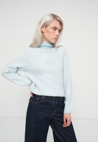 Gaiši zils augstā kakla džemperis ar mīkstu tekstūru un brīvu piegriezumu. Saskaņots ar augstās jostas vietas tumšās džinsa biksēm ar oranžiem šuvju akcentiem.