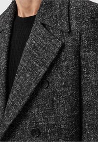 Grå struktureret blazer med dobbeltknap, der har sorte knapper og spidse reverser, parret med et sort strikket plagg underneath.