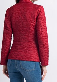 Veste rembourrée rouge avec un col haut, surface texturée, design ajusté et manches longues, associée à un jean bleu.
