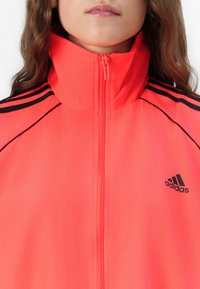 Neonrosa zip-jacka som har en hög krage, svarta axelränder och en broderad svart Adidas-logga på vänster sida.