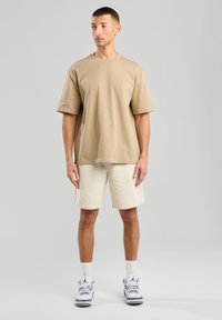 T-shirt in cotone beige a maniche corte, abbinato a pantaloni corti beige chiaro. Presenta una vestibilità rilassata. Indossato con calzini bianchi e scarpe da ginnastica.