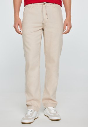 Mees kannab beežeid vabaaja pükse, mis on taljest nööriga, ja valgeid vabaajajalatsid, seisab ühevärvilise tausta ees.