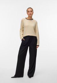 Beige trui met zwarte accenten, ronde hals en lange mouwen, gecombineerd met hoge zwarte broeken met plooien en een tapered pijp.