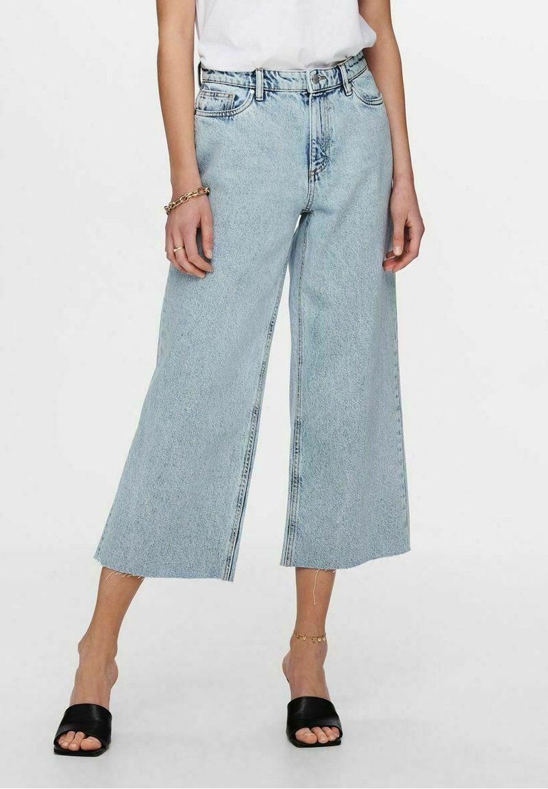 Femme portant un jean large court bleu clair, un haut blanc, des sandales noires à bout ouvert, un bracelet en or et une chaîne de cheville sur fond uni.