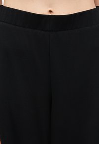 Κοντινή λήψη ενός ατόμου που φοράει μαύρο παντελόνι με ψηλή μέση, εμφανίζοντας τη μέση και την κοιλιά με ορατό τον αφαλό.