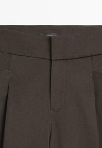 Donkerbruine broek aan de voorkant met een brede tailleband en zichtbare verticale naden, die het ritsgebied toont.