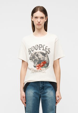 Jeune femme portant un T-shirt blanc avec un motif de panthère rugissante, des fleurs rouges, le texte « KOOPLES » et un jean bicolore bleu, debout droit.