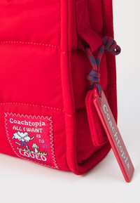Sac en tissu rouge avec un patch cousu présentant du texte et des graphiques colorés. Comprend une étiquette rouge avec "Coachopia" en relief. Surface texturée.
