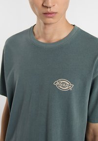 Camiseta de algodón de color verde oscuro con cuello redondo. Presenta un pequeño logo beige en el lado izquierdo del pecho. Mangas cortas y corte relajado.