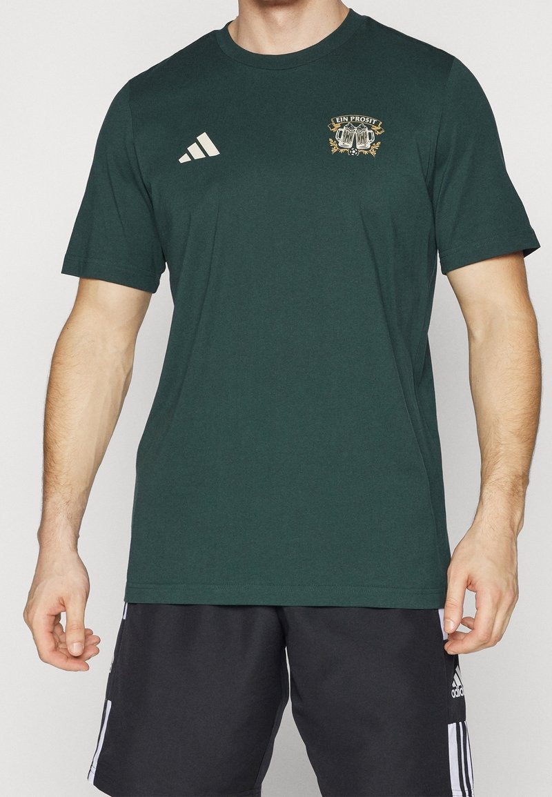 Ciemnozielona bawełniana koszulka z krótkim rękawem, okrągłym dekoltem i złotym emblematem na lewej piersi. Posiada białe paski Adidas na ramieniu.