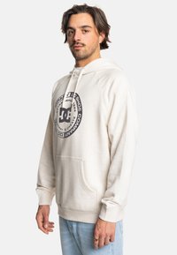 Jasna kremowa bluza z kapturem z kieszenią na przodzie, regulowanym kapturem i okrągłą łatą z ciemnoszarym logo na klatce piersiowej. Miękka tkanina i ściągacze przy mankietach.