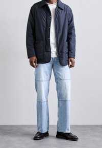 Marinblå vadderad jacka med krage, framfickor, bärs över en vit T-shirt och ljusblå patchwork-jeans med lös passform. Svarta skor.