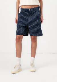 Marineblå shorts med vertikale hvite striper og prikkemønster, laget av et lett stoff med knappelukking og sidelommer.