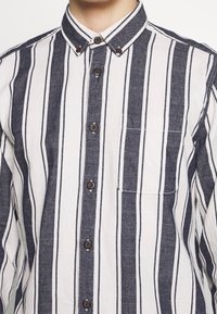 Camicia a righe bianca e blu scuro, con colletto, bottoni marroni e una tasca sul petto con cuciture a contrasto.