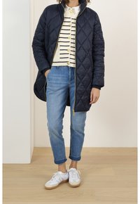 Una donna sta in piedi indossando un cappotto imbottito blu navy, un maglione a righe e jeans chiari, abbinati a sneakers bianche, su un pavimento in legno contro uno sfondo neutro.