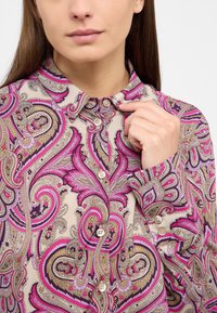 Collared blouse in beige with vibrant pink, purple, and green paisley patterns. Features button closure and long sleeves with buttoned cuffs.