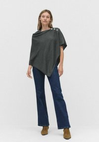 Szary poncho z drapowaną konstrukcją, z trzema dekoracyjnymi guzikami na jednym ramieniu, w zestawie z ciemnoniebieskimi dzwonami i brązowymi botkami za kostkę.