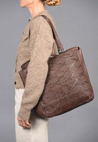 Mujer vestida con un suéter beige y pantalones, llevando un gran bolso de hombro de cuero trenzado marrón sobre un fondo gris.