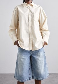 Camisa de botón oversized en beige claro con bolsillo en el pecho, combinada con culottes de mezclilla azul claro con un dobladillo deshilachado y textura desgastada.