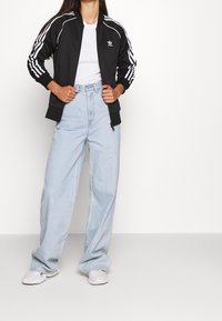 Kobieta w czarnej kurtce dresowej Adidas, gładkiej białej koszulce, jasnoniebieskich szerokich dżinsach i białych sneakersach, stojąca na tle białym.