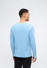 Άνδρας που φοράει ανοιχτό μπλε πουκάμισο με μακριά μανίκια και μαύρο παντελόνι, στέκεται με την πλάτη προς τον θεατή, μπροστά σε μια απλή ανοιχτόχρωμη επιφάνεια.