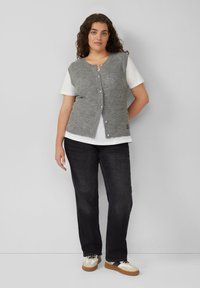 Grijze gebreide vest met knoopsluiting over een wit T-shirt, gecombineerd met zwarte jeans en sneakers met een beige en zwart ontwerp.