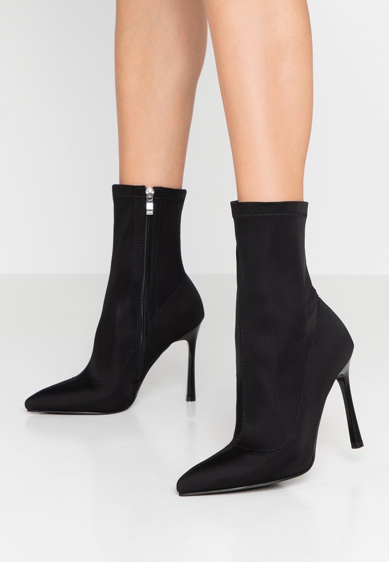 Jambes portant des bottines noires à bout pointu, talons aiguilles fins et fermeture éclair latérale sur un sol blanc.