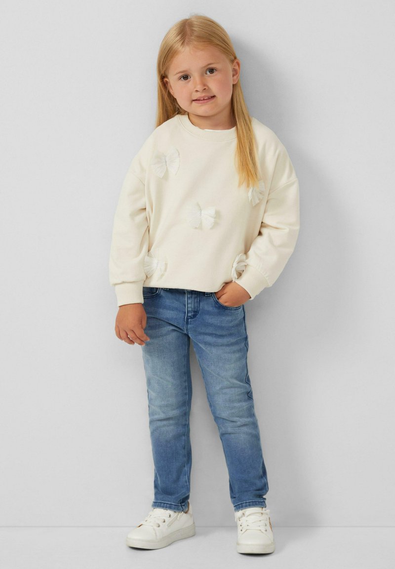 Felpa crema con dettagli a fiocco bianchi, abbinata a jeans di denim blu chiaro. Il modello indossa sneakers bianche, in piedi su uno sfondo neutro.