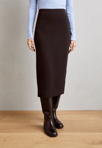 Schwarzer Midirock mit glatter Textur und tailliertem Design, kombiniert mit knielangen braunen Lederstiefeln. Hellblaues, geripptes Langarmoberteil ist sichtbar.