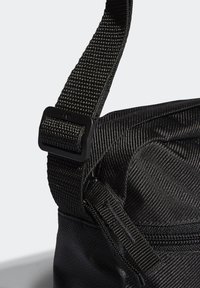 Detail černé nylonové tašky s texturovaným povrchem, nastavitelným popruhem a obdélníkovou plastovou sponou. Zipové zavírání viditelné na boku.