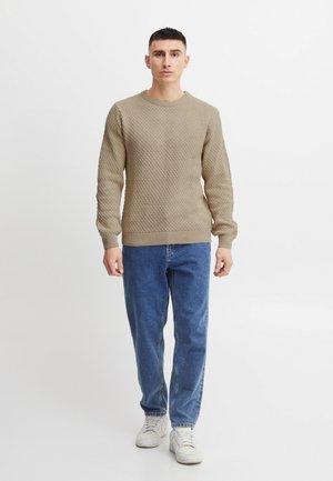 Pull en tricot beige avec motif texturé, col ras du cou, poignets côtelés, associé à un jean en denim bleu et des baskets blanches.