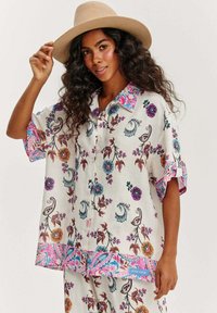 Lněná košile s květinovým vzorem v živých barvách jako modrá, fialová a růžová. Krátké rukávy, zapínání na knoflíky a zakřivený lem.