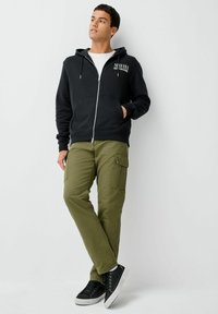 Vyras, vilkintis juodą užtraukiamą džemperį, alyvmedžių žalumo krovinių kelnes, juodus sportbačius, stovi su viena ranka kišenėje prie baltos sienos.