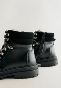 Bottines en cuir noir avec un collier rembourré, éléments en métal, design à lacets et fermeture éclair sur le côté, dotées d'une semelle en caoutchouc texturé.
