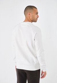 Sudadera blanca de algodón con cuello redondo, mangas largas tipo raglán y puños acanalados. Textura suave y diseño minimalista.