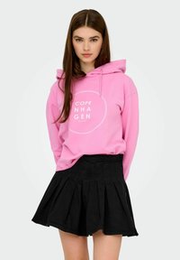 Rosa hoodie med en vit cirkulär grafisk text "COPE COPENHAGEN," matchad med en svart veckad kjol. Mjuk tyg, avslappnad passform.