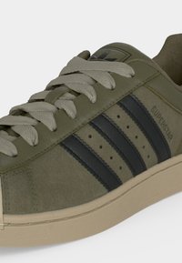 Ολιβ πράσινο sneaker Adidas Superstar με μαύρες ρίγες, μπεζ κορδόνια και μπεζ σόλα, από πλευρική κοντινή γωνία.