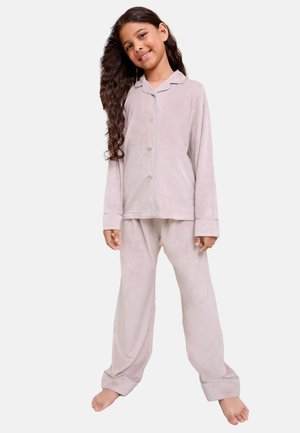 Jeune fille aux longs cheveux bouclés souriante, portant un ensemble de pyjama rose clair à boutons avec manches longues et pantalon, debout pieds nus.
