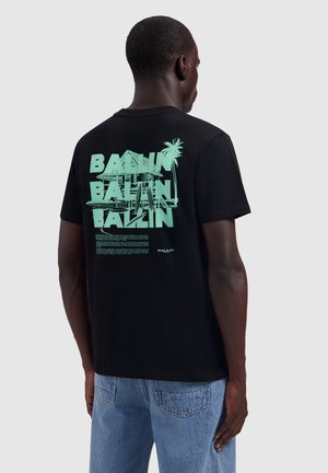 Man draagt een zwart T-shirt met groene grafische tekst "Ballin" en palmboomontwerp op de rug, gecombineerd met een blauwe spijkerbroek.