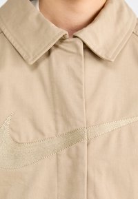 Béžová bunda z tkaniny s velkým vyšívaným zlatým logem Nike na přední straně. Límec je plochý s čistým designem.