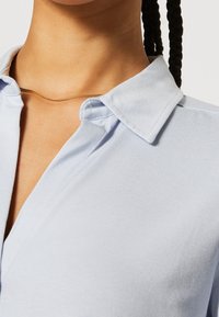 Chemise col bleu clair en tissu doux, avec un devant à boutons et des manches courtes à la texture lisse. Collier en or visible.