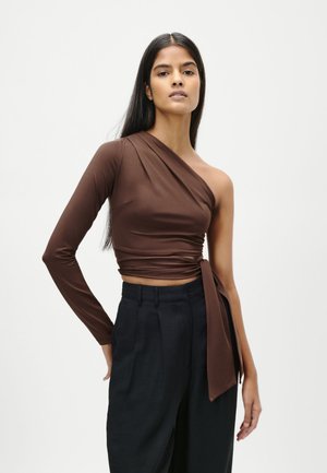 Woman with long dark hair wearing a brown one-shoulder top with a long sleeve and black high-waisted pants against a plain background.