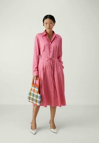 Vestido de camisa rosa de manga larga con cierre de botones y cintura con cordón. La modelo sostiene un bolso a cuadros multicolor. Zapatos blancos de punta.