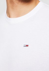 Vit bomullströja med rund hals, slät textur och en liten broderad logotyp i rött, vitt och blått på vänster bröst.