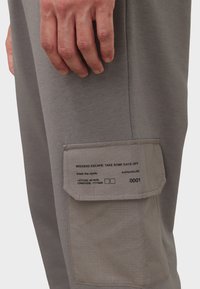 Graue Jogginghose aus weichem Material mit einer seitlichen? Eingrifftasche, die Text und Koordinaten zeigt. Die Tasche hat eine kontrastierende Stofftextur.
