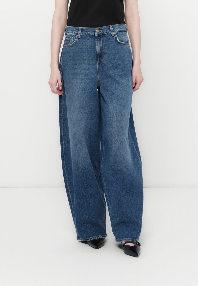 Bredde ben blå denimjeans med høy midje, fem-lomme-design og subtilt falming. Kombinert med svarte spisse hæler.