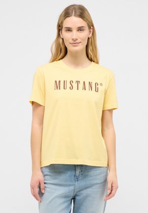 Junge Frau mit langen blonden Haaren, die ein gelbes Mustang-Logo-T-Shirt und hellblaue Jeans vor einem einfarbigen weißen Hintergrund trägt.